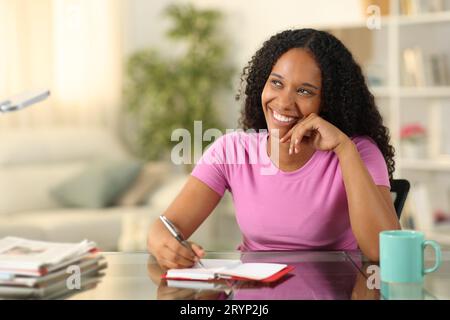 Heureuse femme noire écrivant dans l'ordre du jour rêvant de regarder à côté à la maison Banque D'Images