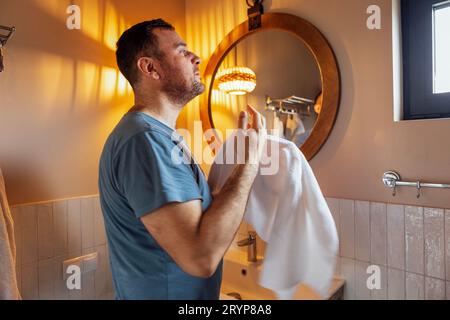 Jeune homme attrayant regarde par la fenêtre et essuie ses mains avec une serviette blanche dans sa salle de bain. Banque D'Images