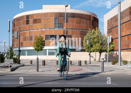 Femme d'affaires élégante en costume vert et casque chevauchant son vélo dans le quartier financier avec un bâtiment moderne sur le fond Banque D'Images