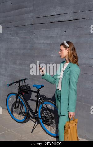 Jeune femme aux cheveux de gingembre responsabilisé en costume vert utilisant smartphone debout avec son vélo sur fond noir Banque D'Images