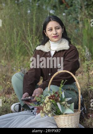 Une belle femme latine assise sur l'herbe sourit dehors au coucher du soleil Banque D'Images