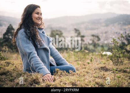 Une belle femme latine assise sur l'herbe sourit dehors au coucher du soleil Banque D'Images