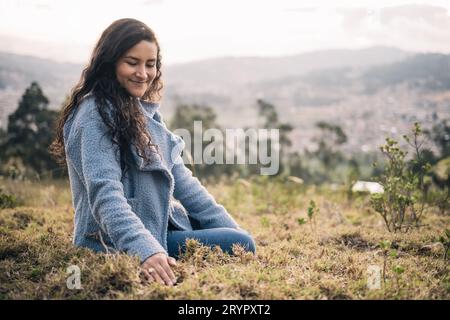 Une belle femme latine assise sur l'herbe sourit dehors au coucher du soleil Banque D'Images