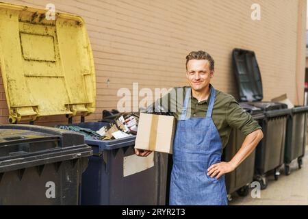 Tri des ordures avec homme souriant blond portant un tablier bleu. L'homme tient une boîte remplie d'ordures près de la poubelle de la ville dans la cour arrière. I. Banque D'Images