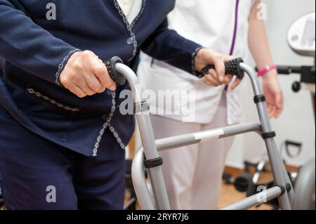 L'infirmière assiste sa patiente âgée sur la marchette pliante. Récupération pour les personnes âgées, soins aux personnes âgées, maison de soins infirmiers. Banque D'Images