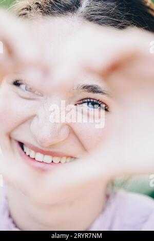 Image rapprochée de femme souriante dans la nature faisant une forme de coeur avec les mains. Femme caucasienne assez joyeuse riant à la caméra Banque D'Images