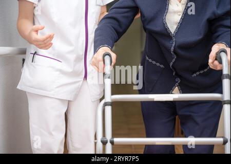 L'infirmière assiste sa patiente âgée sur la marchette pliante. Récupération pour les personnes âgées, soins aux personnes âgées, maison de soins infirmiers. Banque D'Images