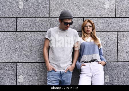 Deux amis penchés sur un mur et ignorant l'un l'autre. Concept de dépendance technologique, gars et une femme en fond de mur gris. Banque D'Images