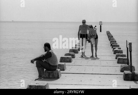 Un observateur solitaire sur une jetée de plage à Bournmouth UK alors que deux nageurs âgés dans leur équipement de natation passent dans cette image en noir et blanc prise en août 1989. Jetée de plage Bournmouth 19890808 Banque D'Images