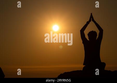 Silhouette du jeune homme faisant du yoga ou de la méditation ou priant au bord d'une montagne au coucher du soleil, Journée mondiale du yoga Banque D'Images