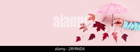Main de femme avec parapluie rose et feuilles tombantes sur fond pastel rose. Concept créatif Hello Autumn Banque D'Images