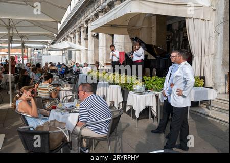 Deux serveurs à manteau blanc près d'un groupe de musiciens se produisent devant des clients assis à Caf Florian sur la Piazza San Marco, (place Saint-Marc) à Venise dans le Venetian Banque D'Images