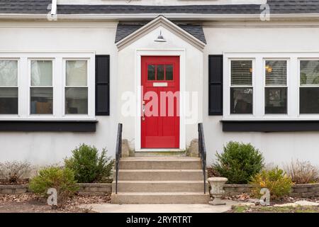 Un détail de porte d'entrée rouge sur une maison blanche avec des marches en béton et des rampes en fer menant à la porte. Banque D'Images