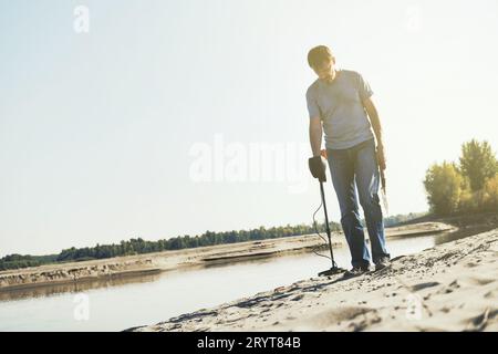 Homme avec détecteur de métaux marche le long de la rive sablonneuse de la rivière. Recherchez des trésors et du métal pour le recyclage Banque D'Images