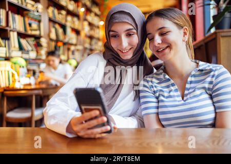 Deux adolescentes de nationalités et de religions différentes sont assises dans la bibliothèque et regardent le téléphone. Des étudiantes charmantes Banque D'Images