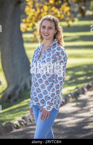 Une jeune femme blonde, habillée de façon décontractée d'une chemise à motifs se tient sous des arbres d'automne dorés Banque D'Images