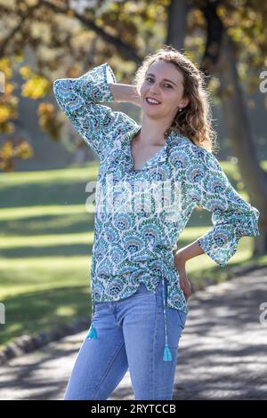 Une jeune femme blonde, habillée d'une chemise à motifs, se tient sous des arbres d'automne dorés avec une main dans ses cheveux et l'autre sur sa hanche Banque D'Images