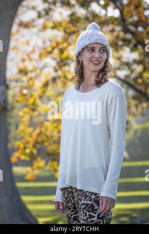 Une jeune femme portant un chandail de couleur crème et un chapeau de laine regarde par-dessus son épaule sous la grande canopée des arbres d'automne Banque D'Images