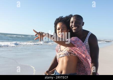 Heureux couple afro-américain embrassant et pointant loin sur la plage ensoleillée par la mer Banque D'Images