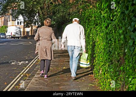 Vue arrière d'un couple âgé marchant dans la rue verdoyante de Londres en automne tenant les mains portant des sacs à provisions Angleterre Royaume-Uni KATHY DEWITT Banque D'Images