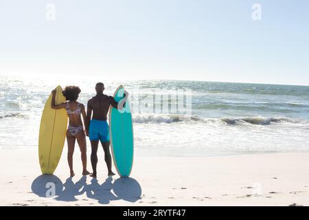 Vue arrière d'un couple afro-américain avec planches de surf debout sur une plage ensoleillée face à la mer, espace copie Banque D'Images