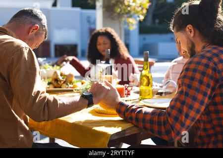 Heureux divers amis masculins et féminins priant avant le repas de célébration de Thanksgiving dans le jardin ensoleillé Banque D'Images