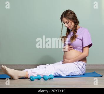 Une femme enceinte est assise sur un tapis de fitness. Femelle regarde son ventre enceinte Banque D'Images