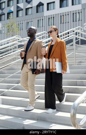 Deux jeunes collègues souriants en vêtements formalwear portant des tasses de café et des gadgets tout en marchant en bas contre l'architecture moderne Banque D'Images