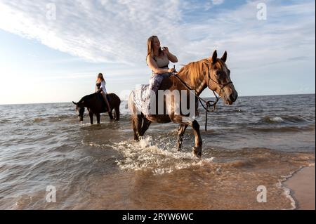 Deux cavaliers féminins à cheval dans le surf d'une plage de l'océan, avec plusieurs autres chevaux en arrière-plan Banque D'Images