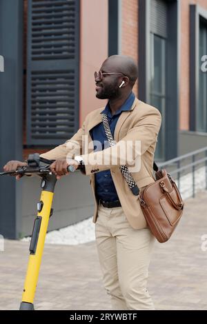 Jeune homme d'affaires prospère en tenue de luxe calme et écouteurs tenant par des poignées de scooter tout en écoutant de la musique dans un environnement urbain Banque D'Images