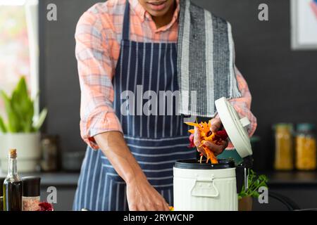 Section médiane de l'homme biracial portant un tablier préparant le repas, compostage des déchets végétaux dans la cuisine Banque D'Images