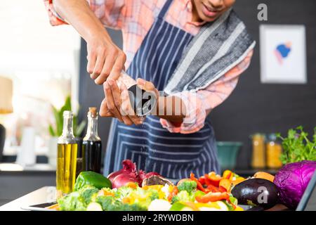 Section médiane de l'homme biracial portant un tablier cuisinant le dîner, assaisonnant les légumes dans la cuisine Banque D'Images