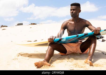 Homme biracial concentré tenant la planche de surf assis sur la plage ensoleillée regardant loin Banque D'Images