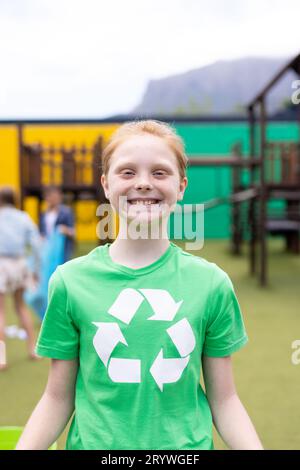 Portrait vertical d'une fille caucasienne souriante portant un t-shirt de recyclage dans la cour d'école, avec espace de copie Banque D'Images