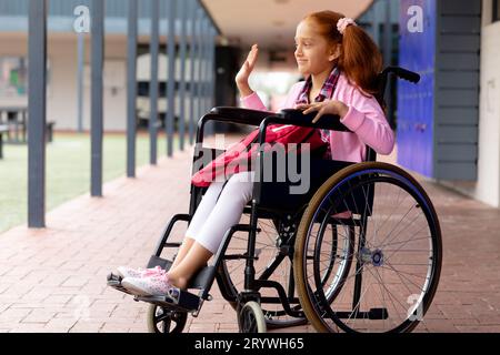 Heureuse écolière biraciale en fauteuil roulant, agitant dans le couloir de l'école Banque D'Images