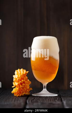 Bière dans une tasse en verre avec une belle mousse épaisse, la mousse coule, une pile de chips de pomme de terre se trouve à proximité sur un fond en bois sombre. Oktoberfest. Banque D'Images