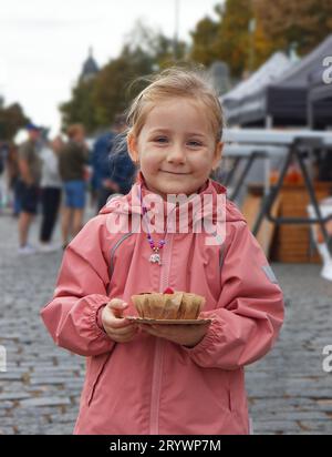 Une petite fille mignonne sourit joyeusement et pose avec le dessert qu'elle vient d'acheter à l'étal sucré un jour d'automne pluvieux au marché des fermiers. Banque D'Images