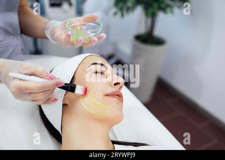 Belle fille caucasienne fait une procédure cosmétique. Le maître de beauté applique un masque rajeunissant sur le visage des clients. Banque D'Images