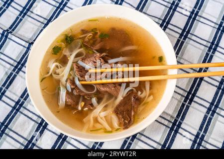 Soupe épicée vietnamienne traditionnelle Pho Bo avec morceau de bœuf, nouilles de riz et herbes dans une assiette avec baguette sur une nappe à carreaux, gros plan. Banque D'Images