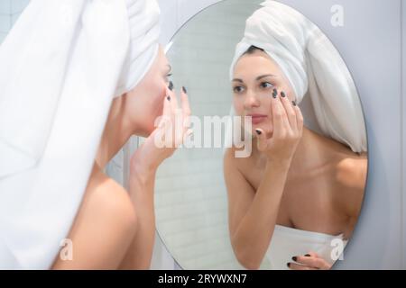 Jeune belle femme appliquant de la crème sur son visage devant le miroir Banque D'Images