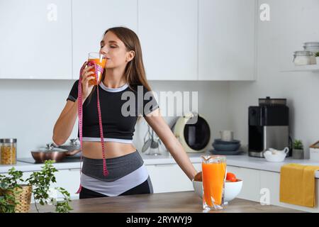 Belle jeune femme avec ruban à mesurer buvant du jus sain dans la cuisine. Concept de perte de poids Banque D'Images