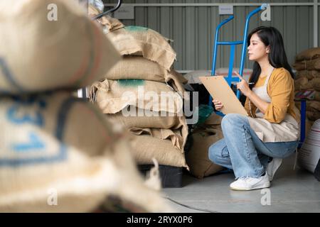 Jeune femme asiatique assise sur le sol dans l'entrepôt de grains de café et vérifiant les documents Banque D'Images