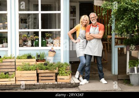 Couple senior souriant portant des tabliers posant à l'extérieur près de leur Greenhouse Bar Banque D'Images