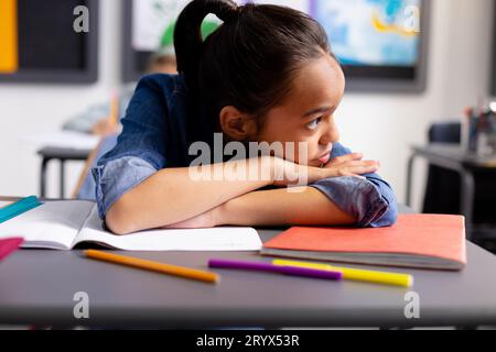 Heureuse écolière biraciale assise au bureau et regardant loin dans la salle de classe de l'école Banque D'Images