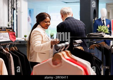 Sympathique souriant femme noire assistante de magasin aidant le client homme senior à choisir des vêtements dans la boutique de mode. Utile agréable vendeuse afro-américaine servant l'acheteur assistant dans le magasin de vêtements Banque D'Images