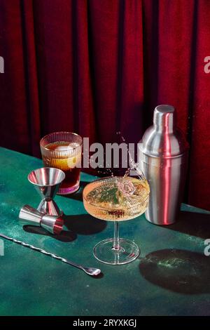 Éclabousser dans un verre à champagne, un verre à cocktail, un shaker, une cuillère à cocktail et un jigger sur une table vert foncé dans une atmosphère de discothèque Banque D'Images