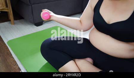 Femme en surpoids s'exerçant pour perdre du poids. Exercice avec des haltères dans des positions d'étirement à la maison dans le salon joyeux Banque D'Images