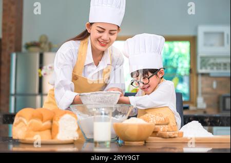 Mignon petit garçon asiatique et belle mère tamisant la farine de pâte avec passoire tamis dans la cuisine à la maison sur la table pour préparer t Banque D'Images