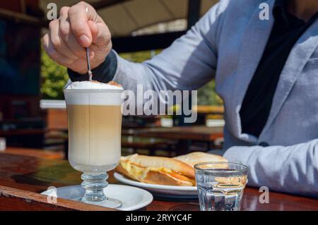 Gros plan homme caucasien irréconciliable assis à l'extérieur du restaurant, mélangeant mousse de cappuccino avec cuillère, concept de nourriture et de boisson, espace de copie. Banque D'Images