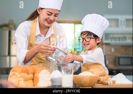 Mignon petit garçon asiatique et belle mère tamisant la farine de pâte avec passoire tamis dans la cuisine à la maison sur la table pour préparer t Banque D'Images